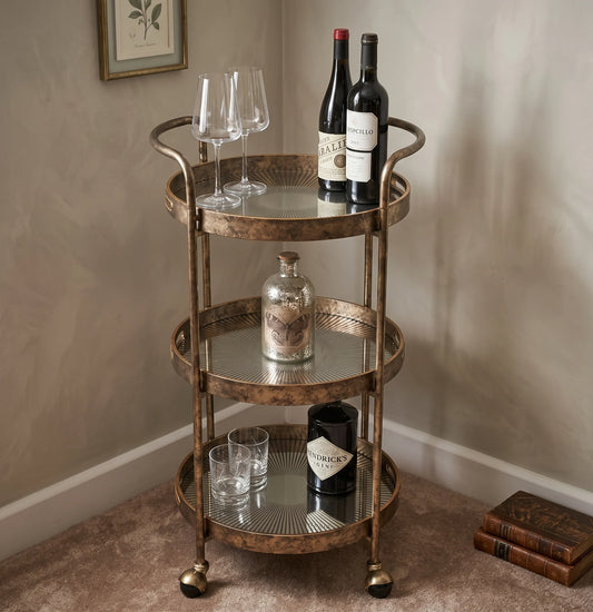 A three-tier rolling bar cart with mirrored glass shelves and a distressed bronze metal frame, styled with wine bottles, glasses, and a decanter in a home bar setting.