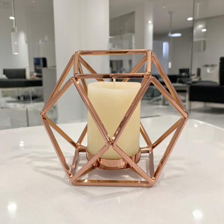 Geometric rose gold candle holder with a white candle on a reflective surface.