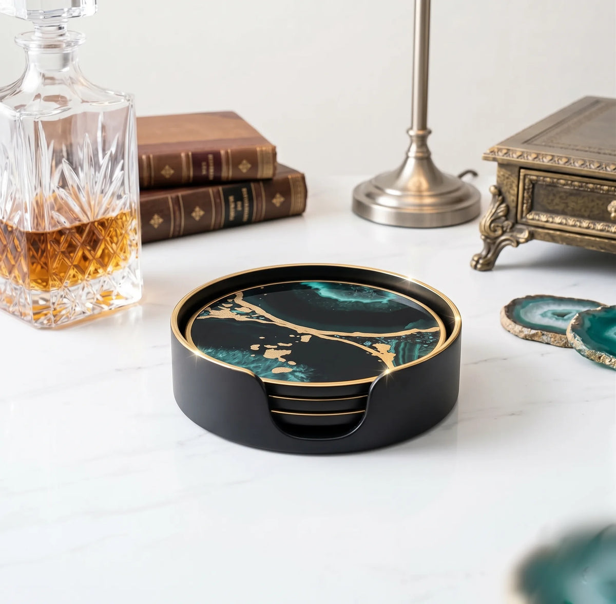A round black coastertainer set with a green and gold marble pattern, displayed on a white marble tabletop with decorative props including a glass decanter, books, and a lamp.