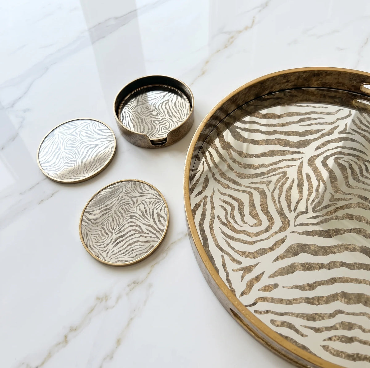 A set of zebra-stripe patterned coasters and a matching serving tray with gold-toned rims, arranged on a white marble surface.