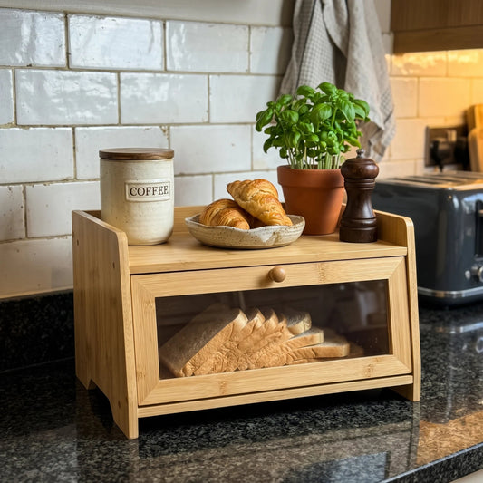 A bamboo bread box with a glass-front lower door sits on a dark kitchen countertop. The box is styled with a ceramic canister labeled COFFEE, croissants in a bowl, a potted basil plant, and a wooden pepper mill.