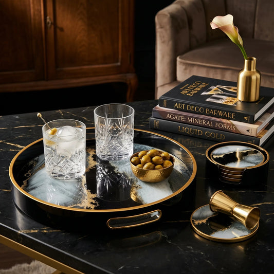 A luxury bar accessories set displayed on a black marble table. The set includes a round black tray with gold trim and a white marbled center, a matching small coffee table, a gold jigger, a gold cocktail spoon, and a gold olive holder.