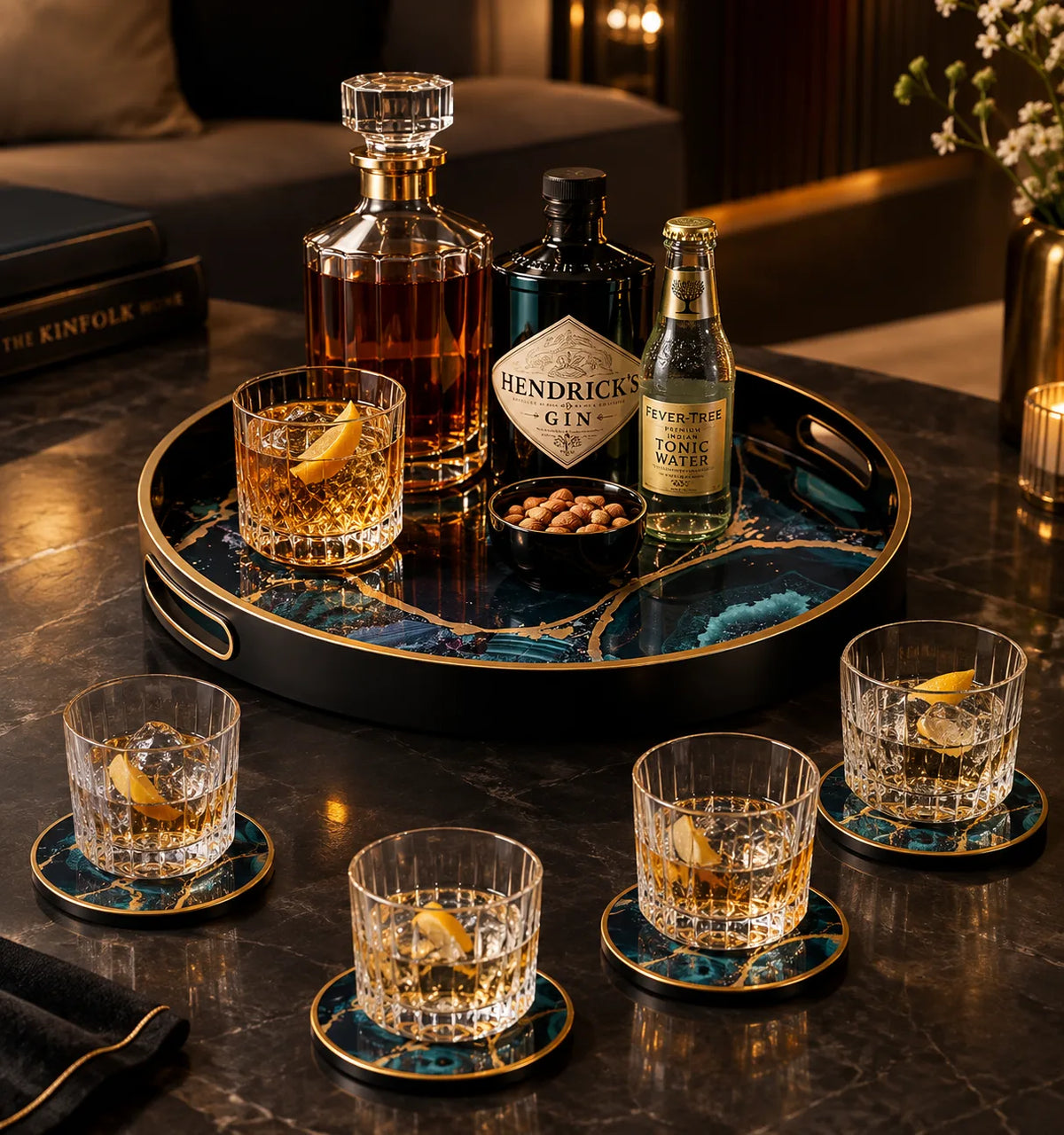 A luxury bar scene with a round black and teal marble serving tray holding a whiskey decanter, Hendricks Gin bottle, King Collins soda bottle, two rocks glasses with lemon slices, and a small bowl of mace. Four matching rocks glasses with coasters are arranged around the tray on a dark marble table in a dimly lit lounge setting.