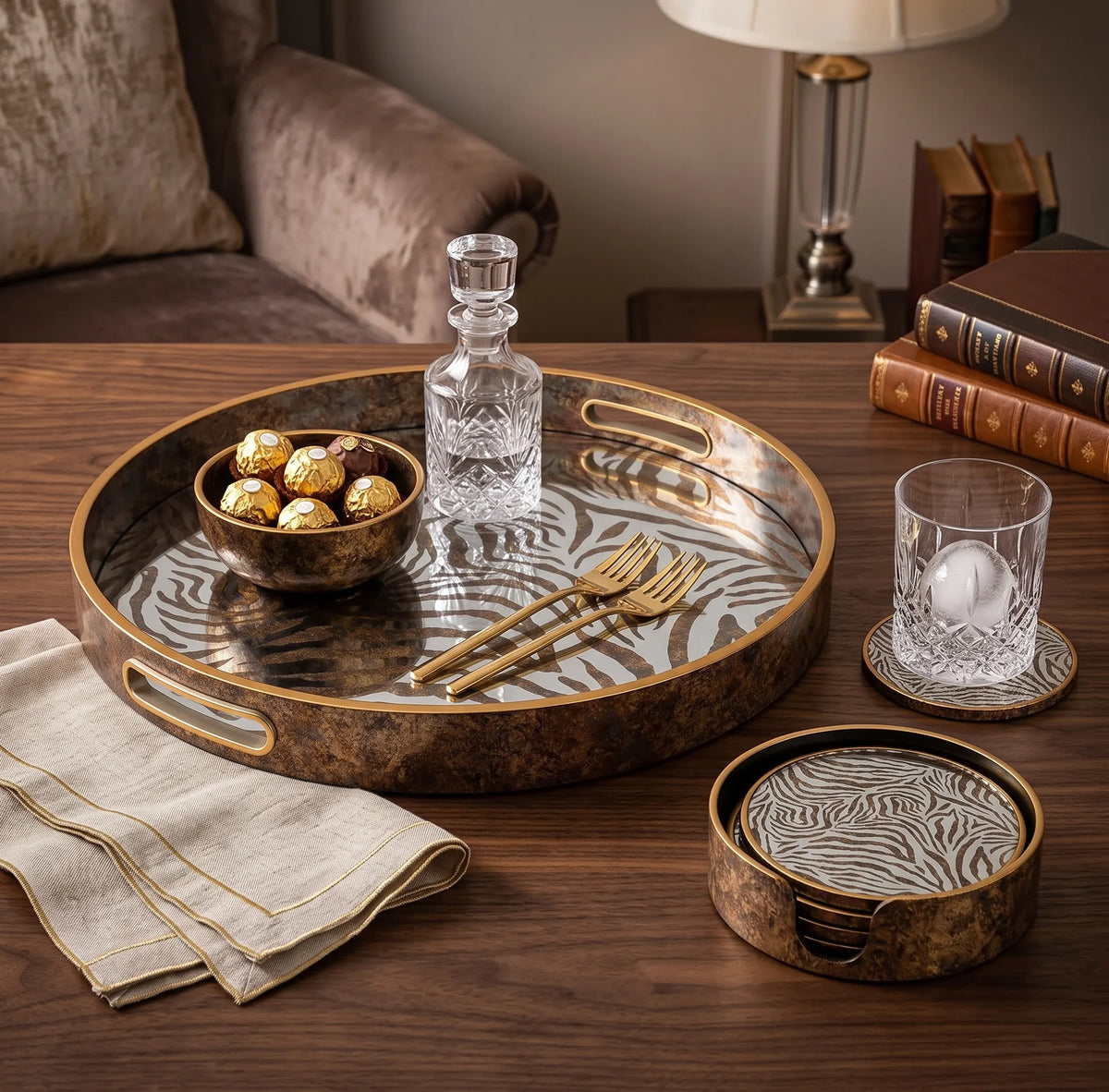 A round decorative tray set in a warm, styled living room setting. The set includes a large oval tray with a zebra-stripe pattern on a bronze-brown background, a matching small round tray, a matching glass coaster, a clear glass decanter, two gold-toned forks, and a glass of water on coasters.