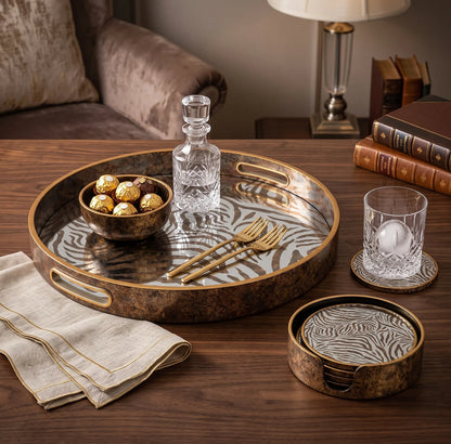 A round decorative tray set in a warm, styled living room setting. The set includes a large oval tray with a zebra-stripe pattern on a bronze-brown background, a matching small round tray, a matching glass coaster, a clear glass decanter, two gold-toned forks, and a glass of water on coasters.