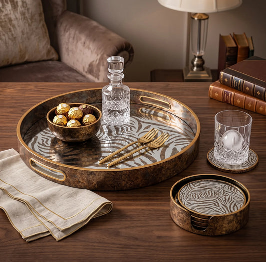 A round decorative tray set in a warm, styled living room setting. The set includes a large oval tray with a zebra-stripe pattern on a bronze-brown background, a matching small round tray, a matching glass coaster, a clear glass decanter, two gold-toned forks, and a glass of water on coasters.