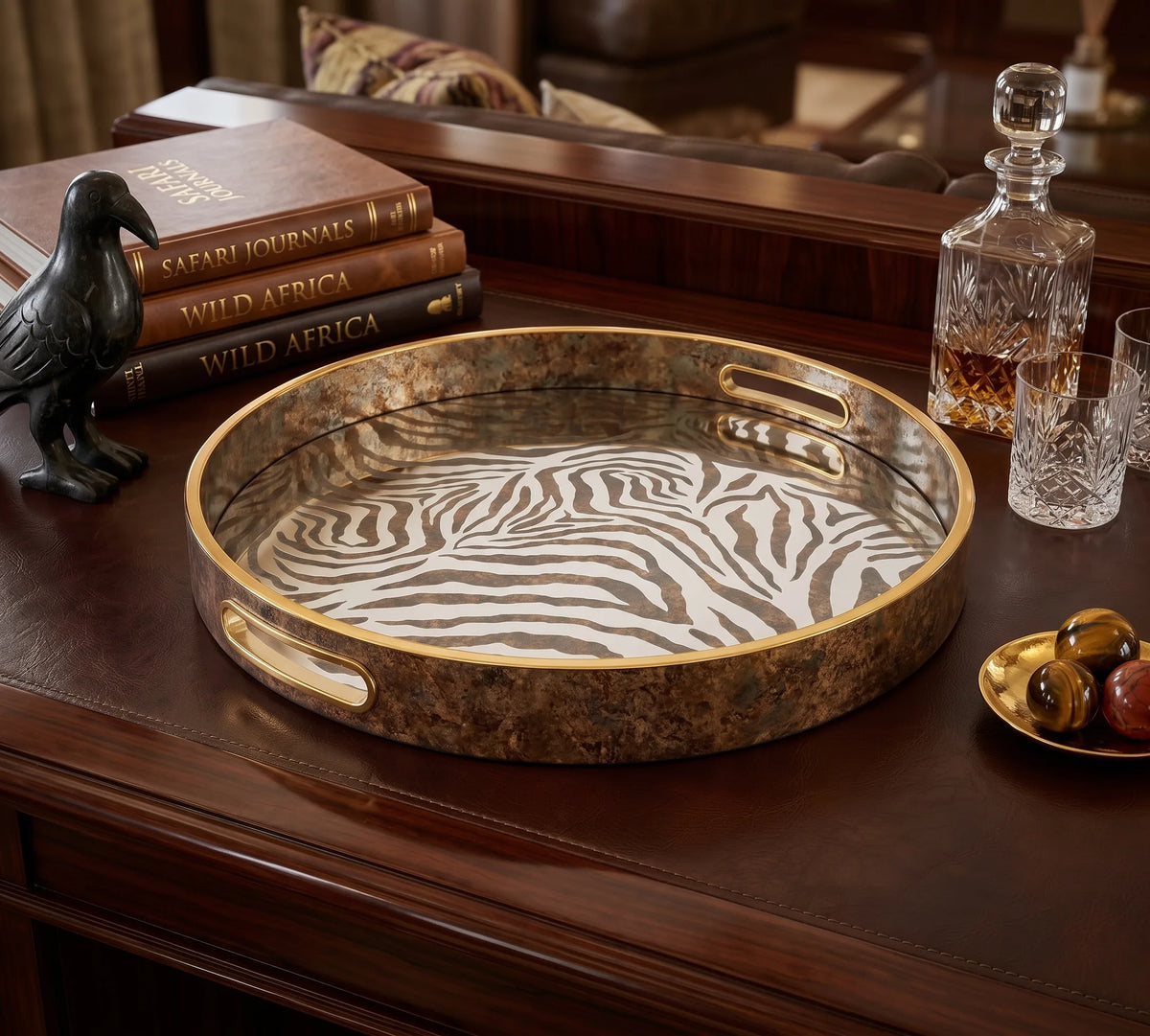 A round decorative serving tray with a zebra-stripe pattern on a dark brown background, featuring gold-toned metal handles and a gold rim, displayed on a dark wood table with books, a raven figurine, and glassware.
