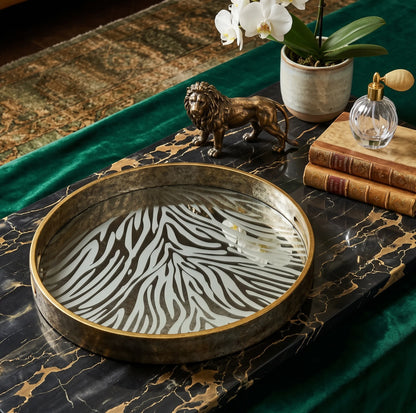 Round decorative tray with a zebra-stripe pattern on a dark marble surface, styled with a bronze lion figurine, books, a glass perfume bottle, and a potted orchid.