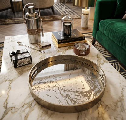 A round mirrored tray with a brushed gold metal frame sits on a white marble coffee table in a styled living room setting.